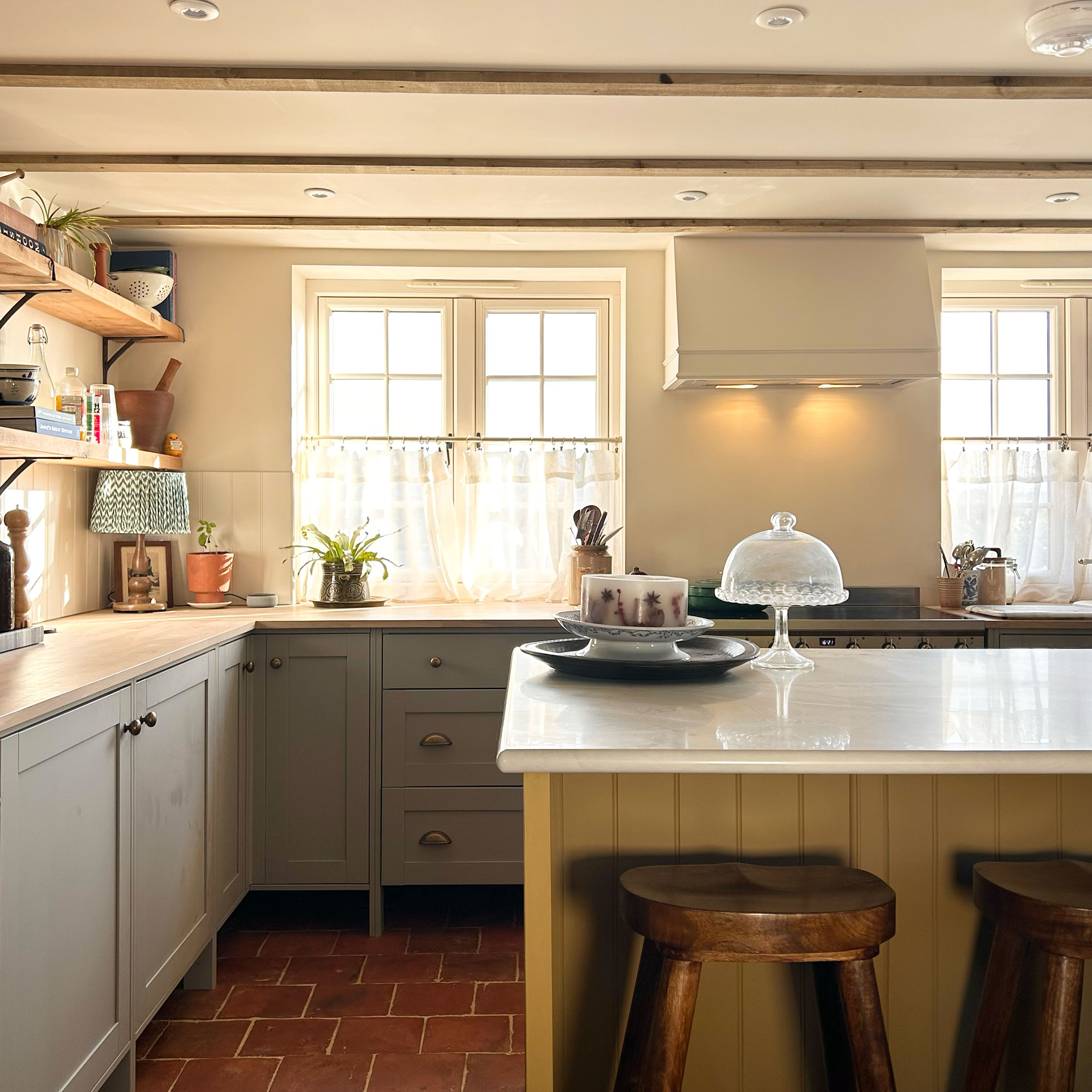 Country kitchen with terracotta tiled flooring, island with yellow panelling and dark wooden bar stools, pale blue-grey cabinetry and slimline ceiling beams