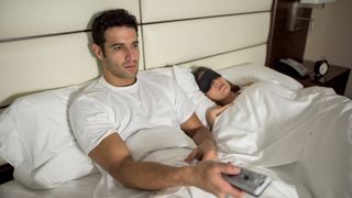 Man sat up in bed using a remote control on a TV with his partner asleep next to him with an eye mask on