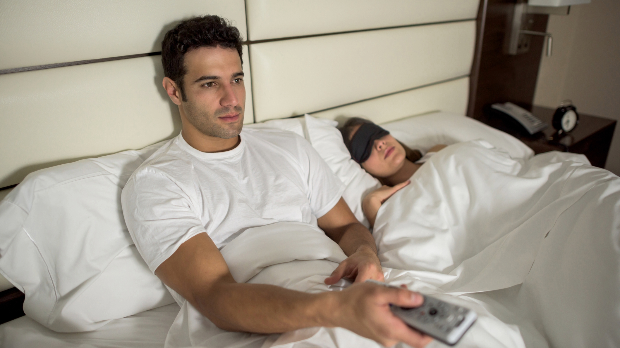 Man sat up in bed using a remote control on a TV with his partner asleep next to him with an eye mask on