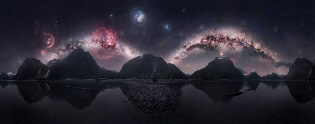 Person stands on a rocky shore before reflective water, jagged mountains, and a vivid star-filled galaxy sky
