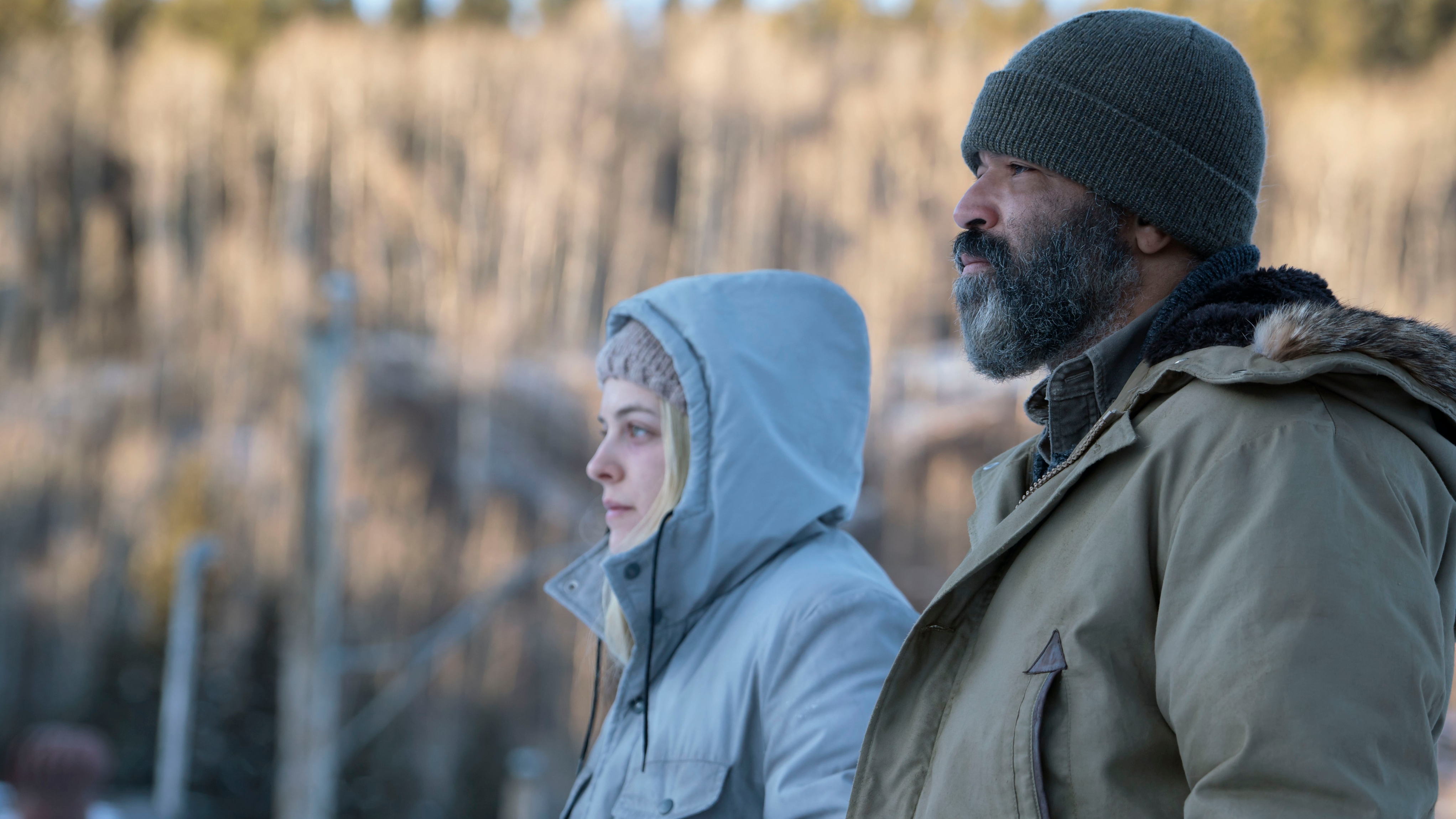 Hold the Dark image of two people in thick coats looking away from the camera and surrounded by trees and snow.