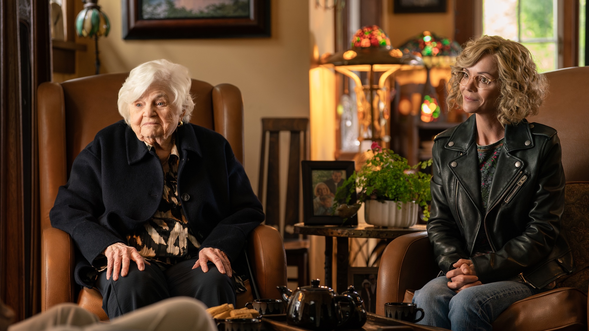 June Squibb's Geraldine wearing black coat sitting in a tan chair next to Christina Ricci's Misty wearing her leather jacket and jeans in front of a tea set and snacks in Yellowjackets Season 4