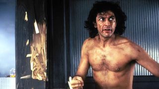 A young Jeff Goldblum stands shirtless next to a broken wall