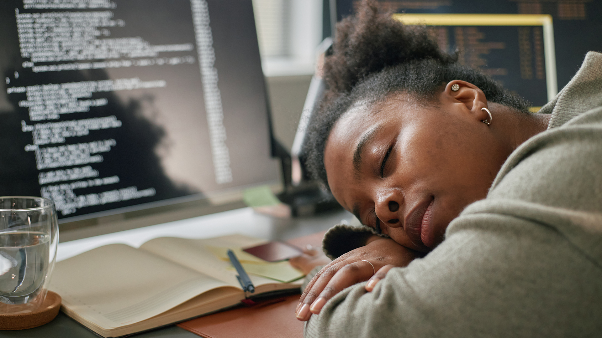Female software developer sleeping with head placed on desk, with computer monitor with code in background and open notepad on desk.