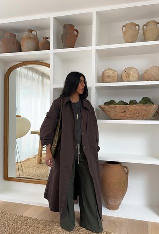 a photo showing the best summer-to-fall transition items with a woman standing against shelves wearing a brown trench coat over a gray sweater with green trousers and a green bag