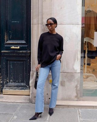 Die Mode-Influencerin Sylvie Mus posiert auf einem Pariser Bürgersteig und trägt eine ovale Sonnenbrille, einen schwarzen Pullover mit Rundhalsausschnitt, eine schwarze Mini-Tasche, gerade Jeans, schwarze Strumpfhosen und schwarze Pumps