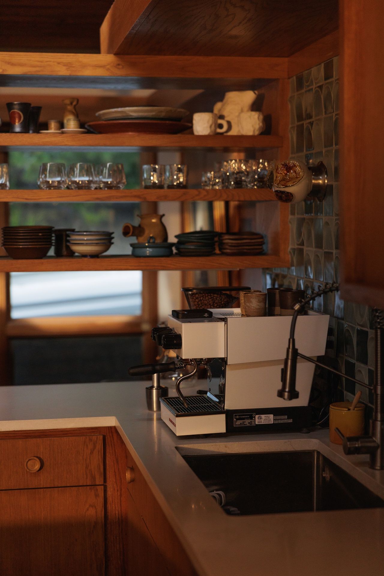 a white La Marzocco coffee machine on a white countertop in a wooden kitchen with open shelving that is full of different tableware and glassware.