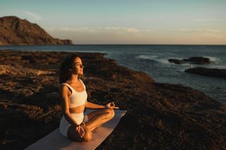 Quiet meditation for mental health. Sportive woman sitting in lotus position on yoga mat on the beach. Female wellbeing and life balance, physical activity for good health. Beautiful warm sunlight at sunrise, morning awakening.