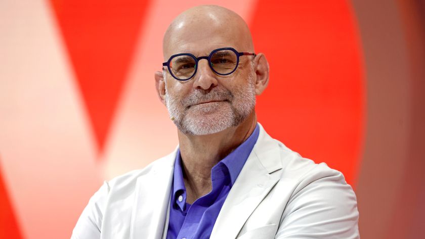 Bestselling Novelist, writer, Executive Producer, Harlan Coben in a purple shirt and white suit jacket sitting in front of a red background and smiling for the camera 