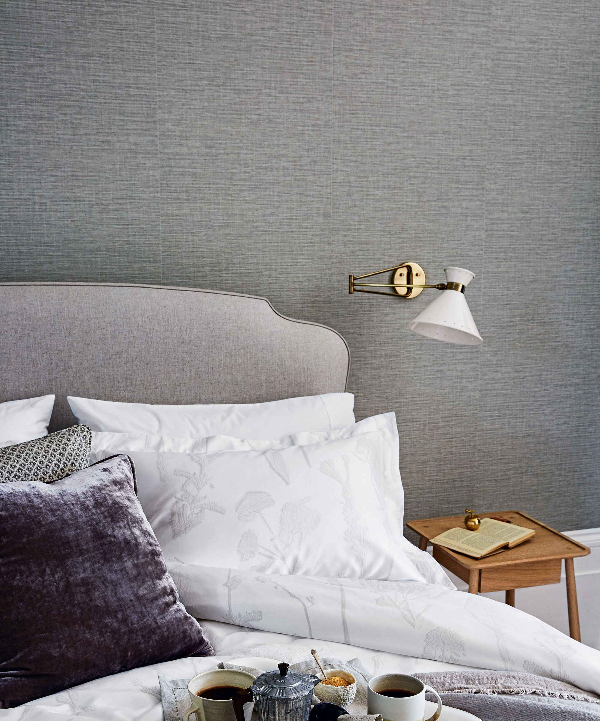 A gray bedroom with a white bedside wall lamp and a bed with a gray upholstered headboard