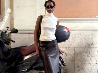 Paris-based fashion influencer Sylvie Mus poses on a street in Rome, Italy, in front of a peach-colored wall and a motorbike, wearing black cat-eye sunglasses, a white sleeveless turtleneck, a shoulder bag and a leather skirt