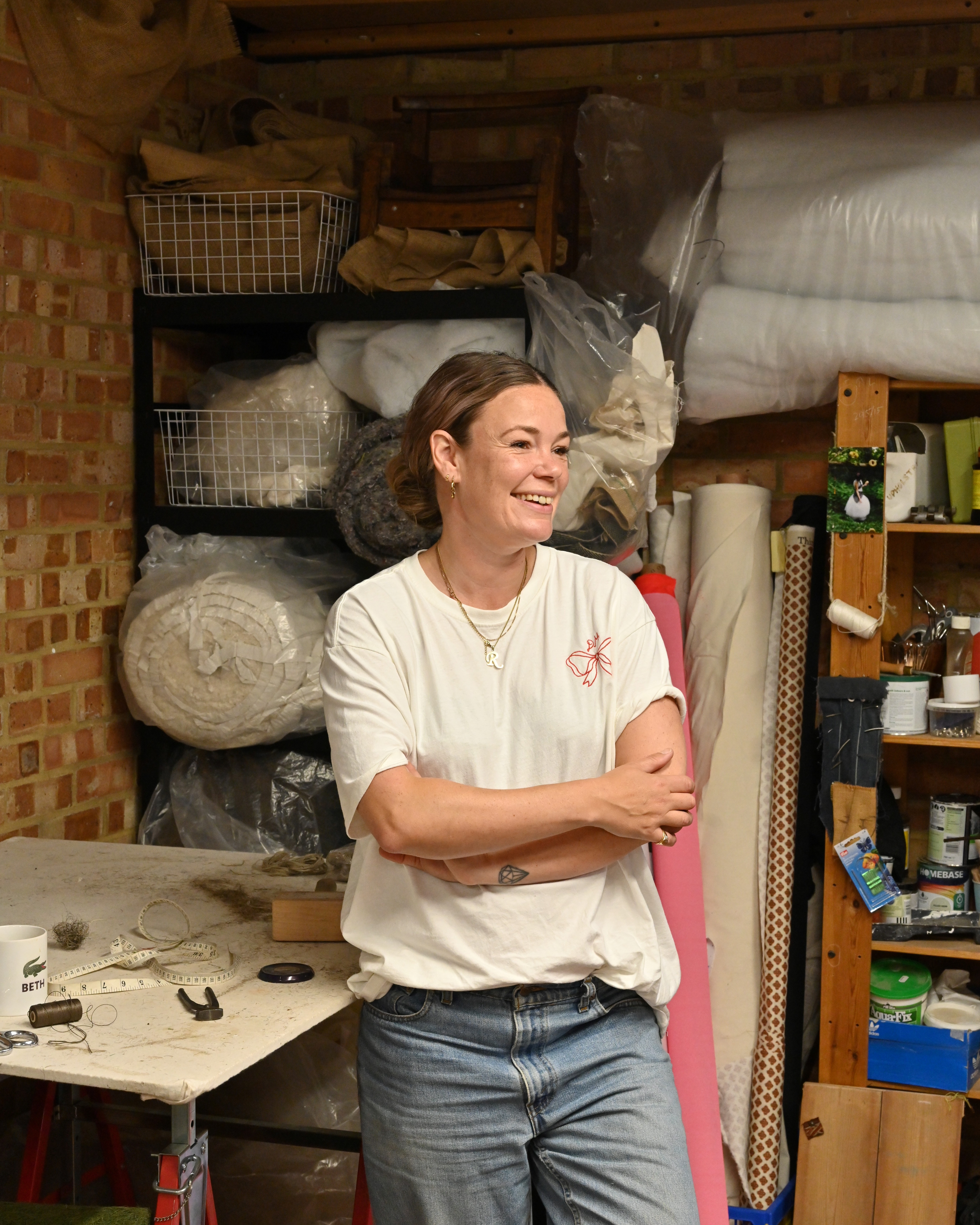 A female upholsterer photographed at work, up close and in portraits, in her studio, filled with wooden furniture, colorful fabric rolls, and mockup designs, and illuminated by bright light.