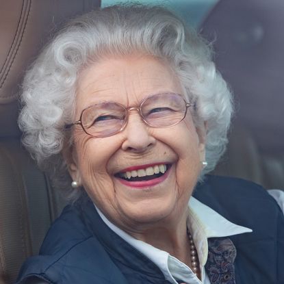 Queen Elizabeth wearing a blue vest and white shirt laughing while driving her car 
