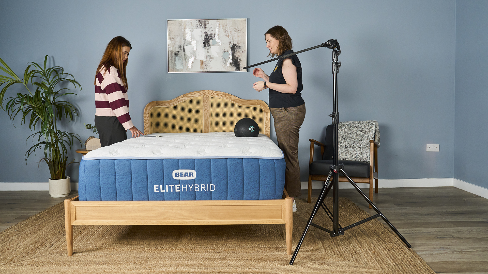 A full shot of a sleep studio where two mattress testers are standing on either side of the Bear Elite Hybrid mattress. A large black exercise ball has just been dropped on the right hand side of the bed and the tester on the other side is using an accelerometer to measure the motion transfer across the bed. A tall, black pole is positioned to the right of the bed, indicating the height the ball was dropped from.