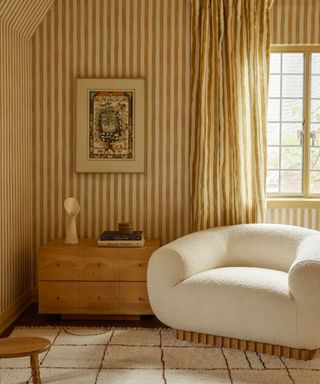 lulu and georgia striped wallpaper in a corner of a bedroom with matching ochre curtains, a boucle armchair and a vignette with a sideboard art and decor