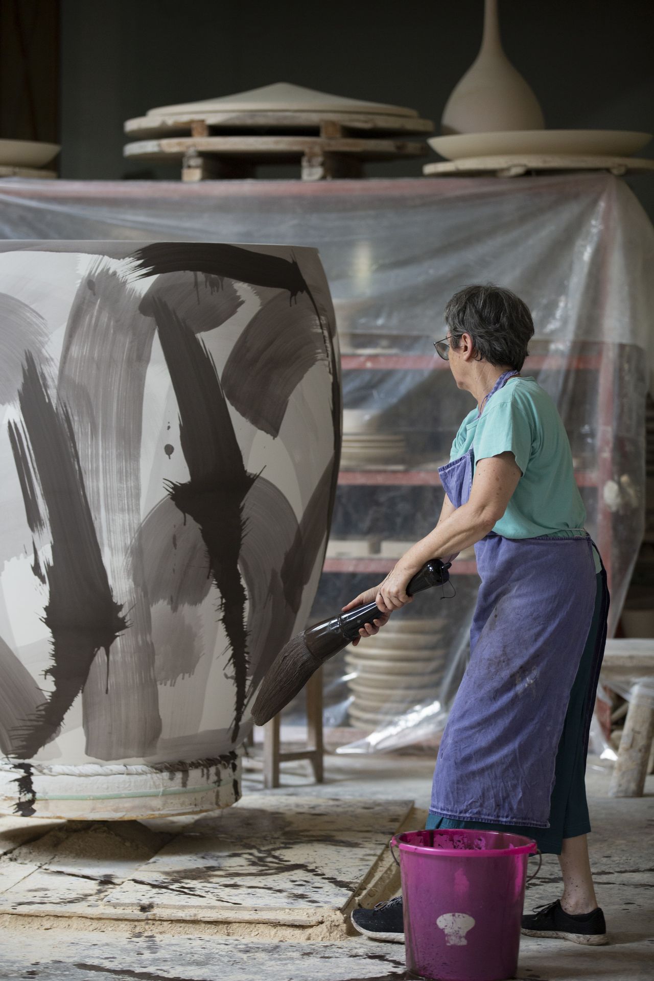 Felicity Aylieff, dressed in denim and a blue top, works and observes her large-scale porcelain vases.