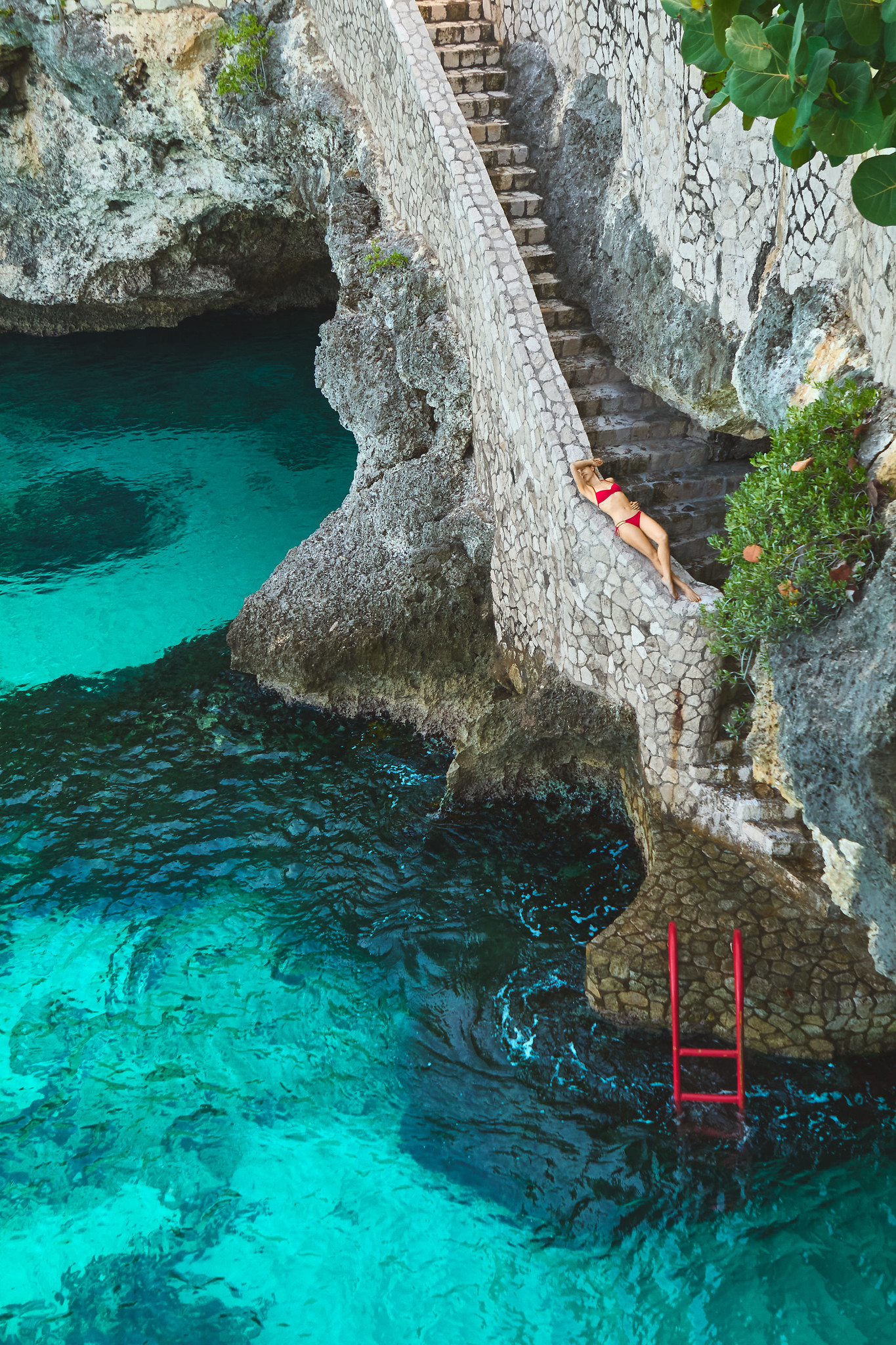 Stone steps leading down to the sea at the Rockhouse Hotel in Jamaica. A red ladder is pictured that dips into the water. A woman in a red bikini is lounging on the stones.