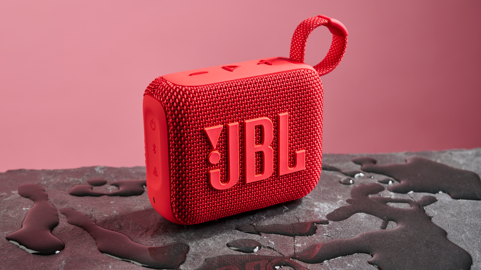 A red JBL Go 4 pictured against a pink background. It is sitting on a dark stone surface with puddles of water around it.