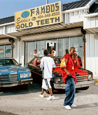The front of Eddie’s Gold Teeth/Famous Eddie’s shop with men standing outside in front of two cars.