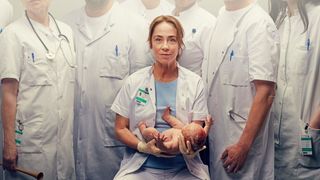 Sofie Gråbøl i kittel sidder med et nyfødt barn og er omgivet af mænd i kitler