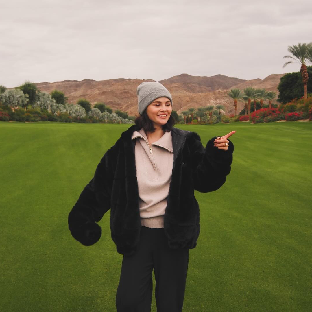 Selena Gomez wearing a beanie and black jacket, standing in a field