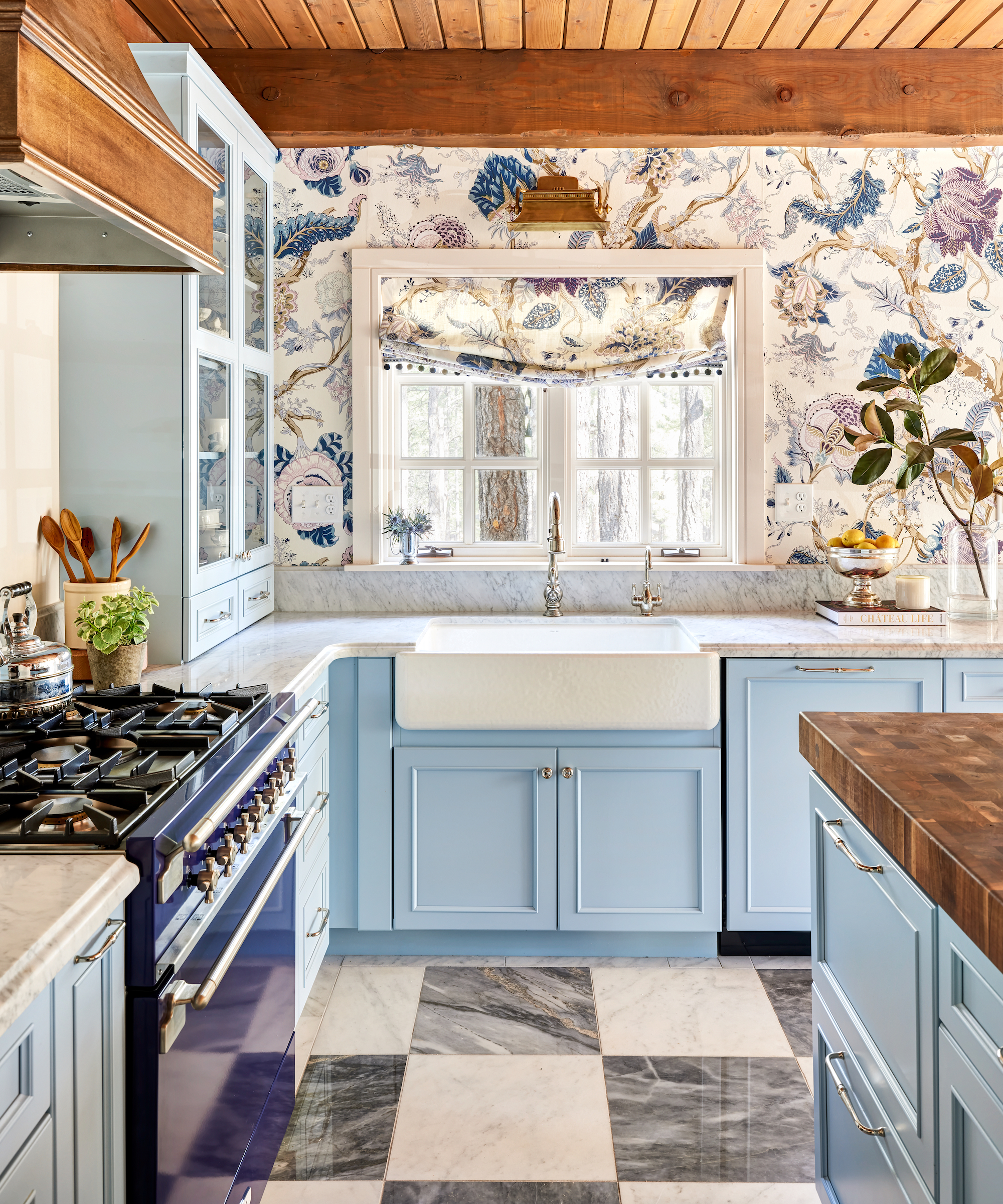 Kitchen with pale blue units, range with wooden hood, butler sink, patterned wallpaper and patterned blind
