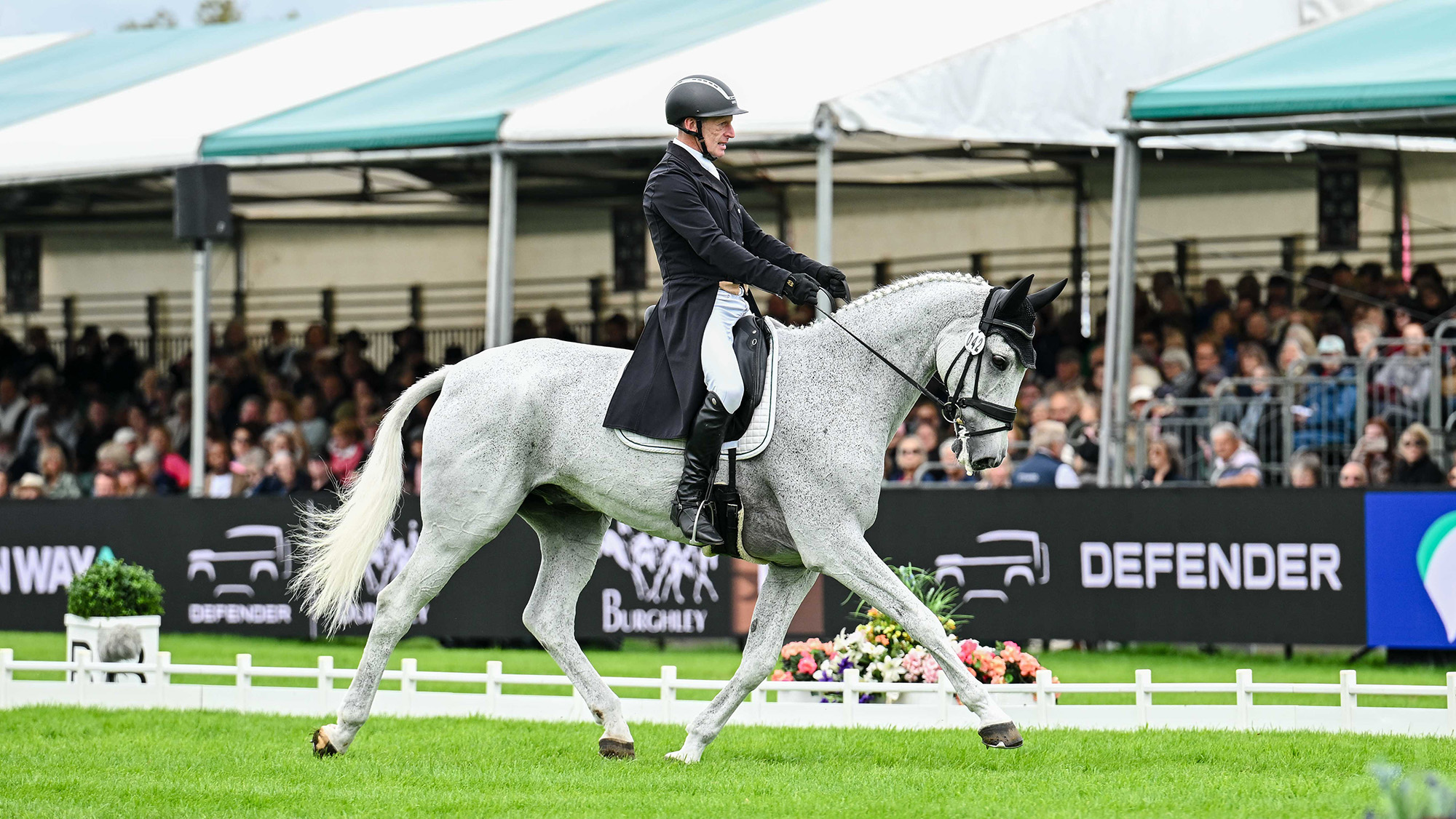 Anthony Clark, a rider in a black tailcoat, rides grey horse Ballinaguilkey Fortunus in the grass dressage arena at Burghley