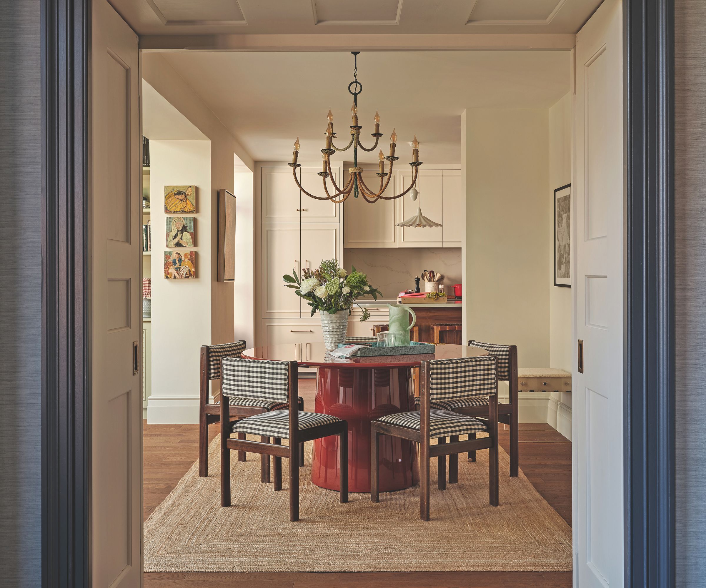 Dining room with a round red table, black-and-white checked chairs, a brass chandelier, and a view into the kitchen beyond