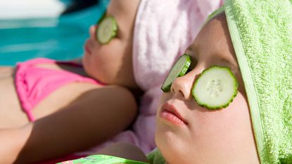 Two little girls lounge by a pool with their hair wrapped in turbans and cucumber slices on their eyes.