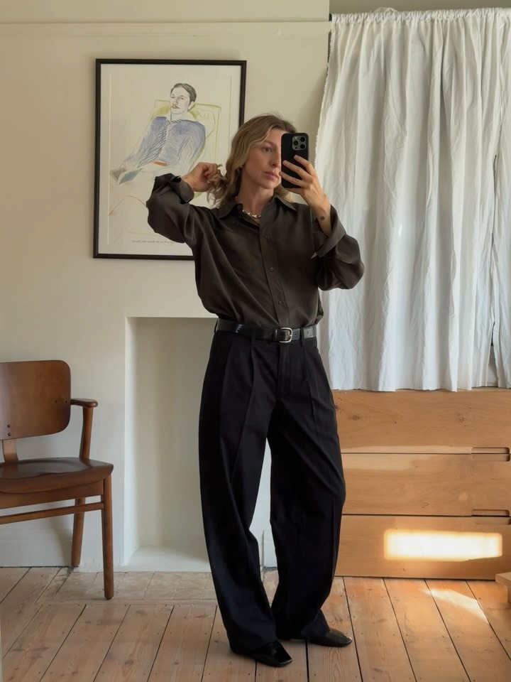 Woman takes a mirror selfie wearing pleated wide leg trousers, a brown button down and black belt.
