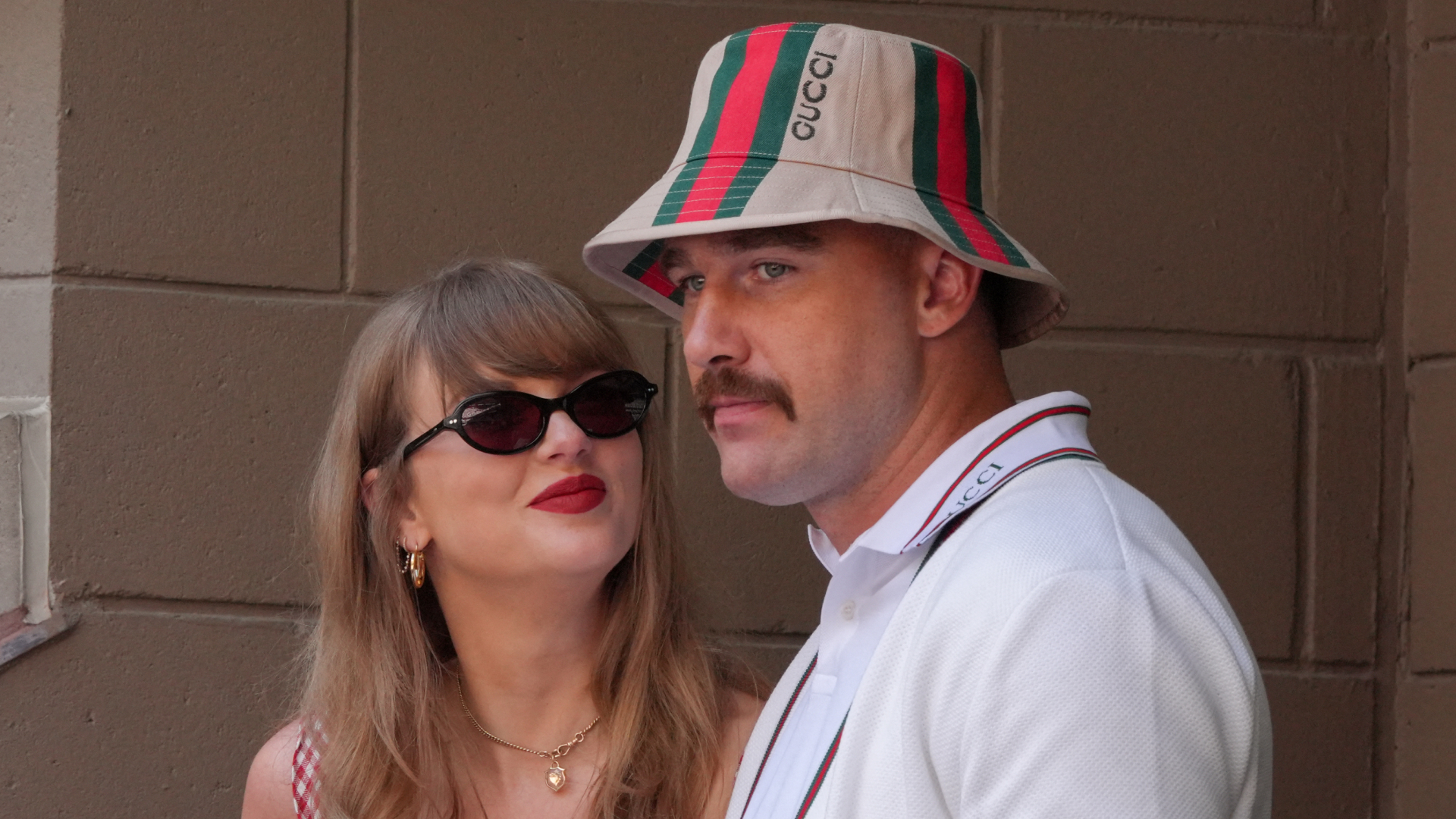 Taylor Swift wears a red gingham dress and black sunglasses and smiles up at boyfriend Travis Kelce, who is wearing a white polo shirt and Gucci bucket hat
