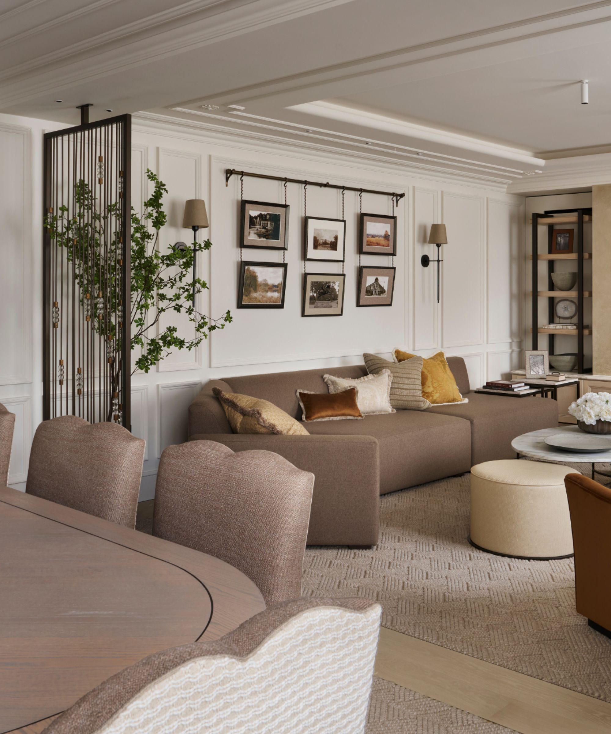 Neutral, cozy living room with brown sofa and scatter pillows, coffee table and ottoman in front gallery wall and lights on wall above, greenery, shelves in the background, dining chairs in foreground
