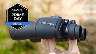 The Celestron Cometron 7x50 binoculars being held by a hand with a green and brown background with a black Space.com Prime Day deals badge in the upper-left corner.