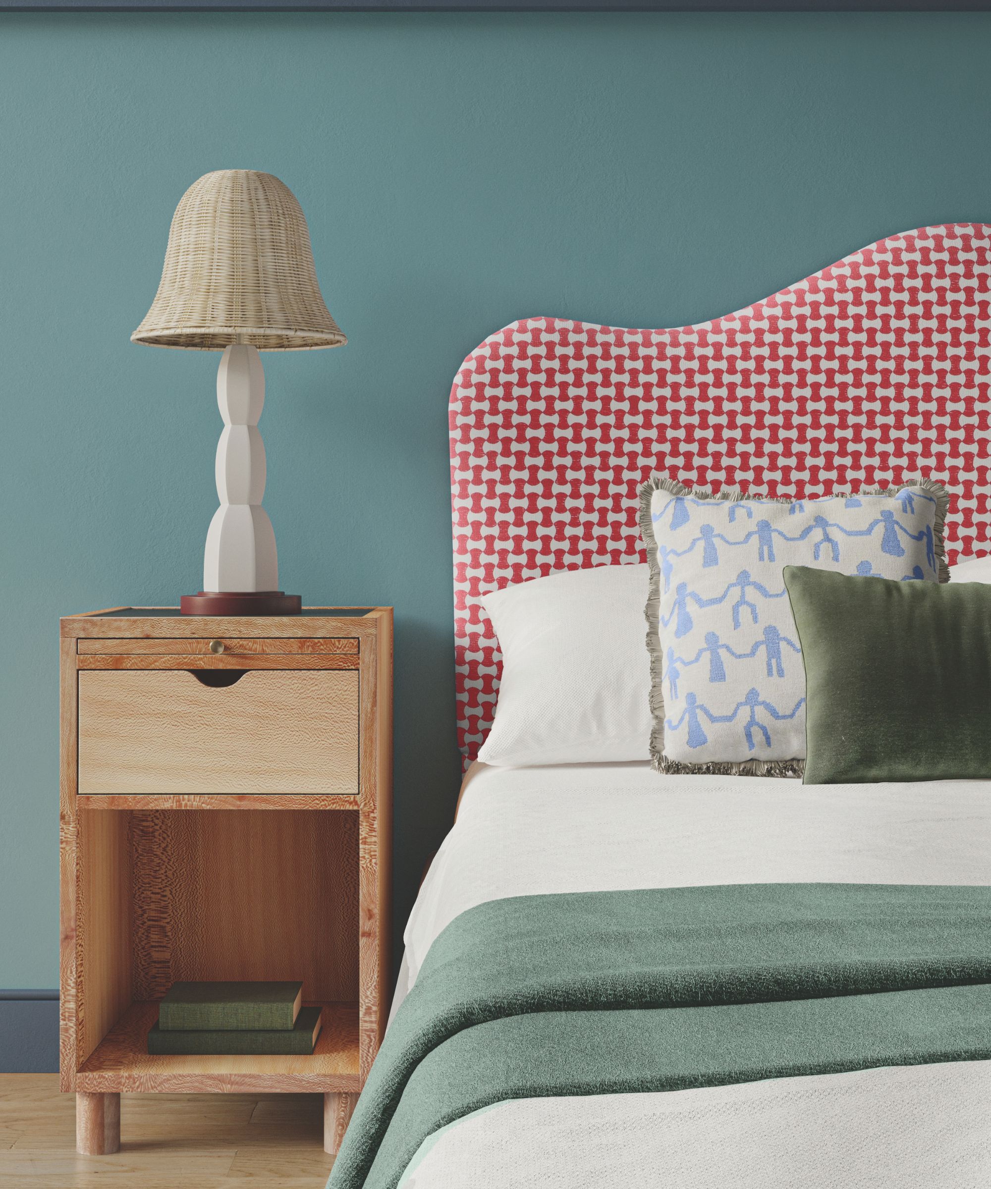 A blue bedroom with a double bed with red patterned headboard, white bedding, a patterned cushion and green velvet cushion, blue blanket, and blue walls. Beside the bed, to the left, is a wooden beside table with a white lamp with rattan lampshade, and the floor is light wood.