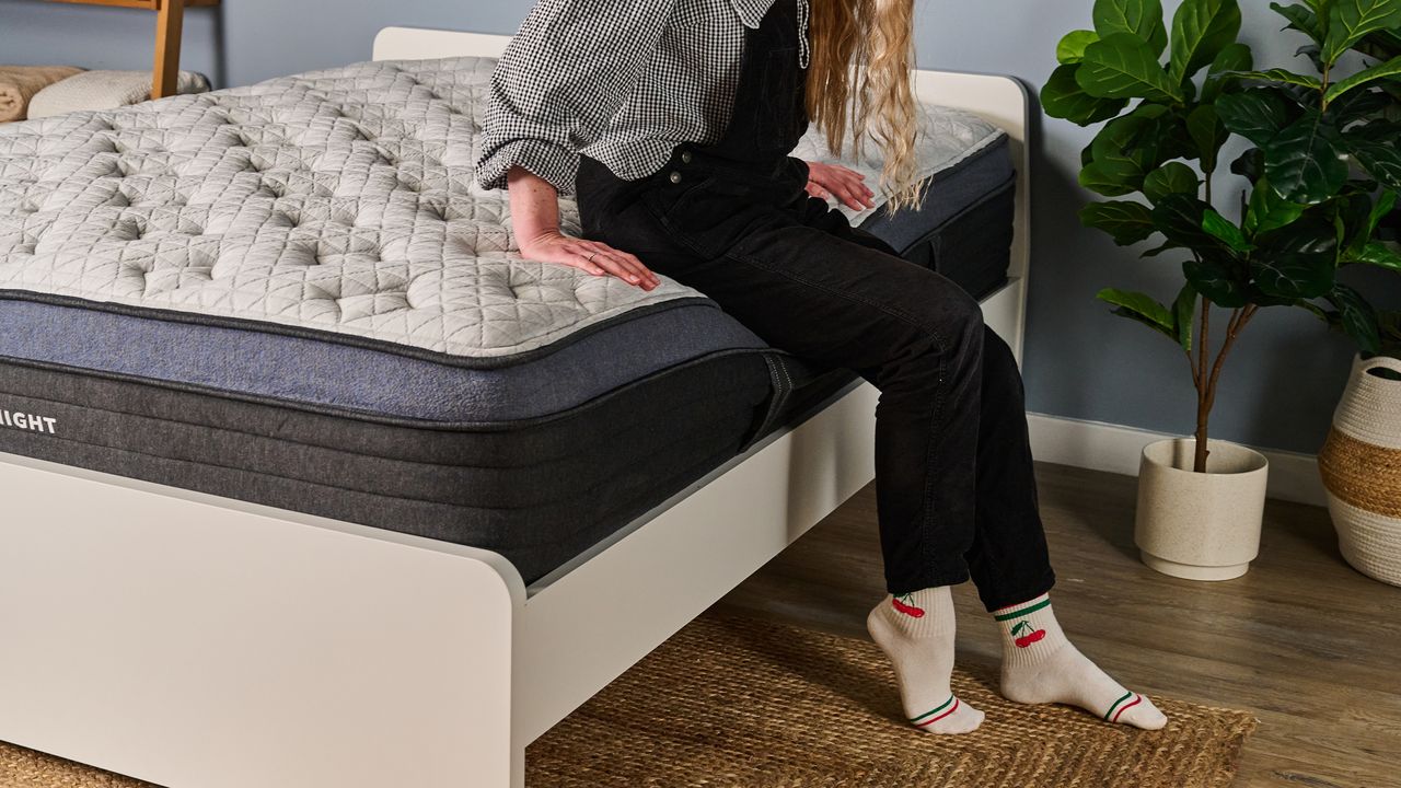 The image shows a woman sat on the edge of a white mattress with navy blue trim, placed on a white bed frame in a pale blue bedroom