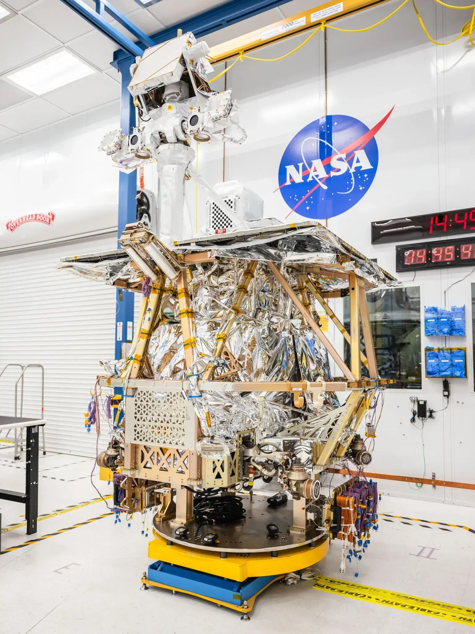 A foil-wrapped robotic explorer is stowed on a yellow mounting pad. a brass-pipe-looking frame of supports circles the body in a hexagonal pattern, with a rectangular shelf resting flat above. A mast in the top middle front supports an apparatus. The rover stands in a white room with a white floor and white ceiling. A NASA logo is painted on the wall in the background, above mounted data displays with red numbers.