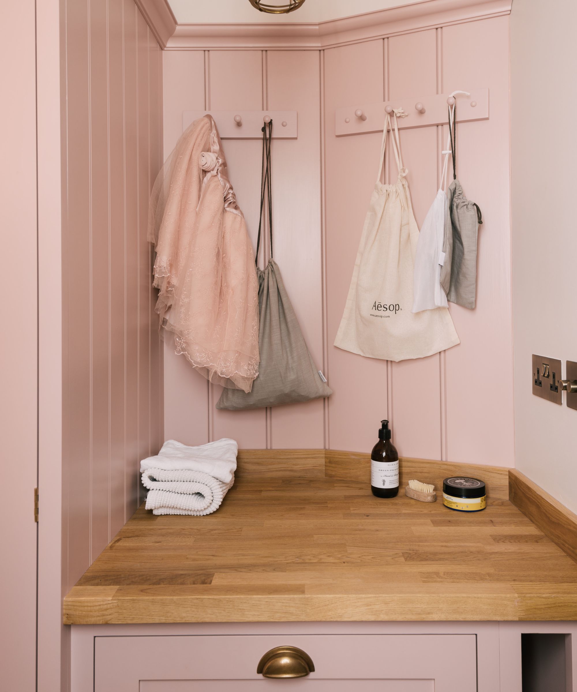 A small corner utility or boot room area painted in blush pink, featuring shaker-style panelling with wooden peg rails for hanging bags and garments. A solid oak countertop provides a warm contrast, accessorised with neatly folded towels and a few grooming essentials.