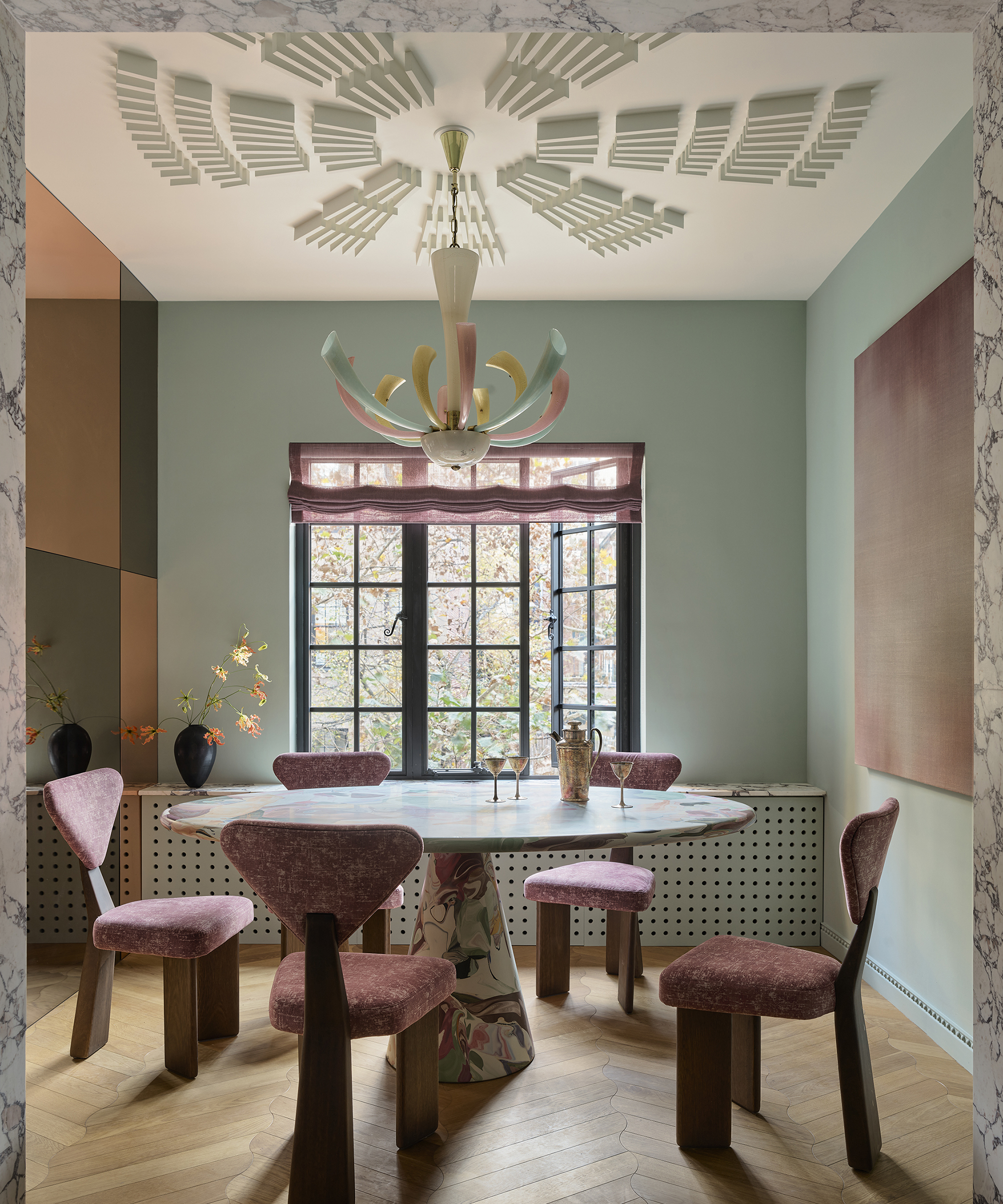 A pale green-blue dining room with a sunburst decal on the ceiling and pink dining chairs around a round marbled pedestal table