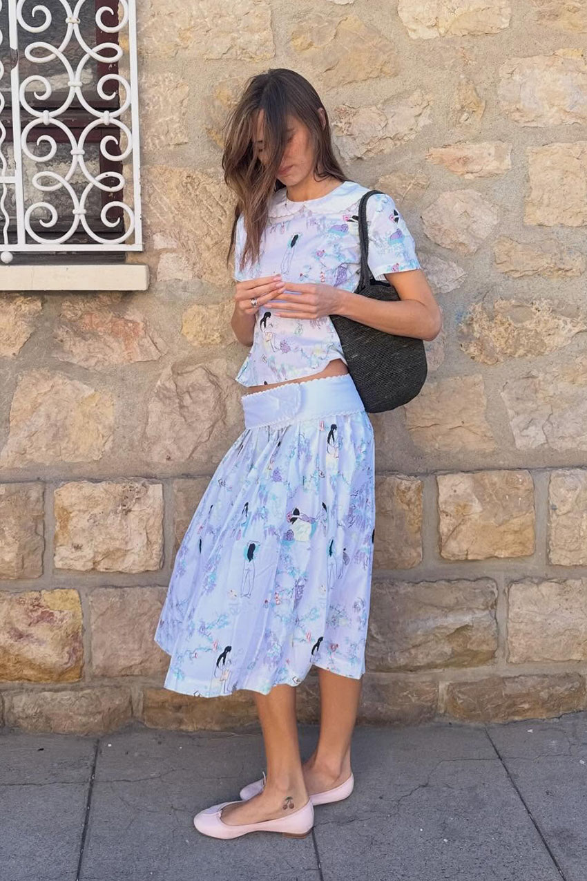 A woman wearing a white printed Peter Pan&ndash;collar top with a matching A-line skirt, a brown woven bag, and light pink flats.