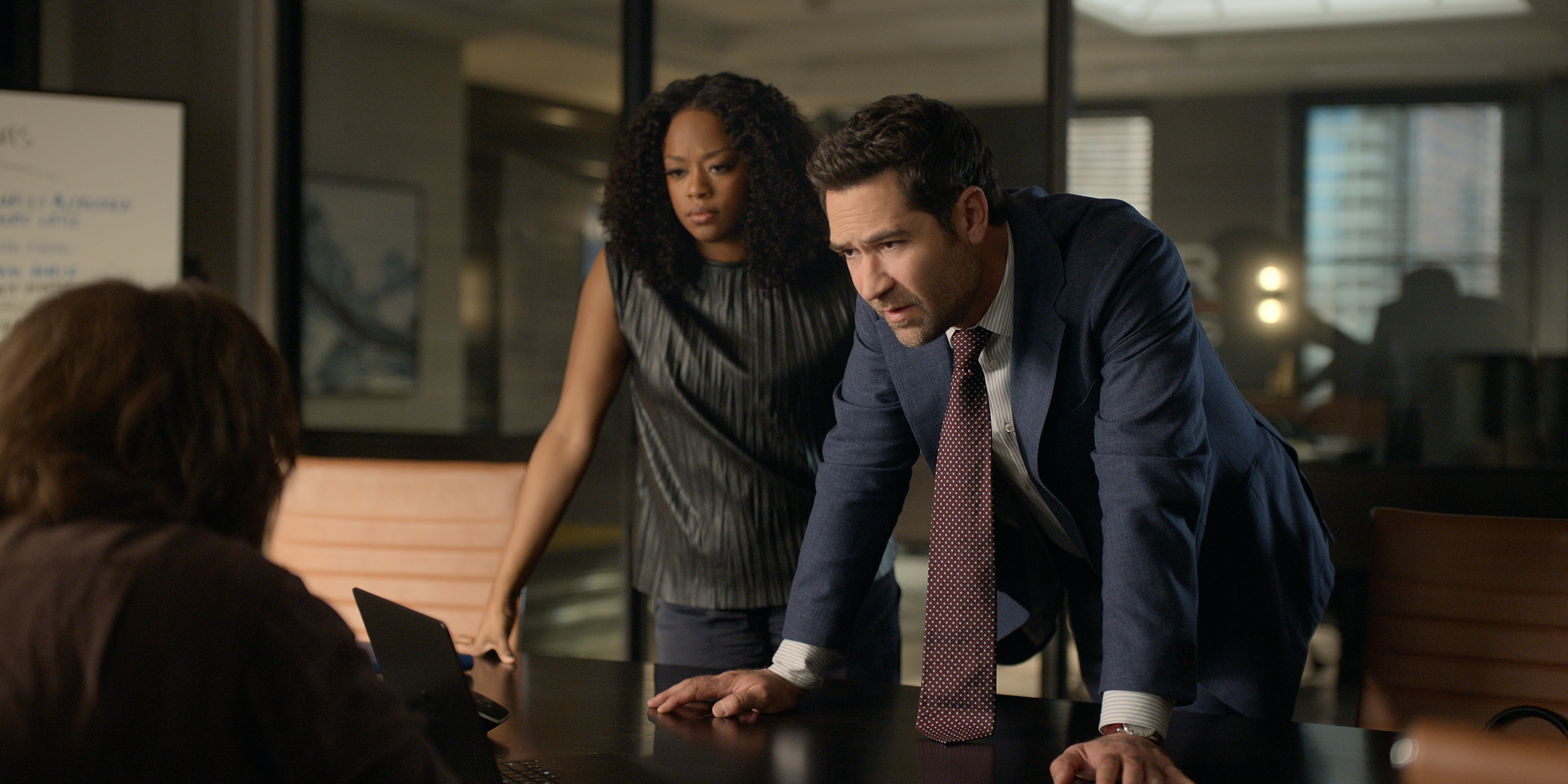 Jazz Raycole as Izzy Letts and Manuel Garcia-Rulfo as Mickey Haller leaning over a table to talk to someone in an office in episode 404 of The Lincoln Lawyer