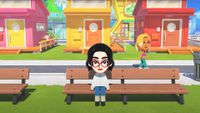 Screenshot from the reveal trailer of Tomodachi Life: Living the Dream, showing a female Mii sat on a bench as another female Mii walks on the street behind her.