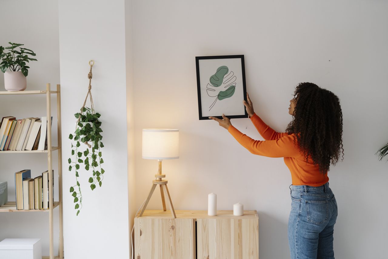 A woman hanging a photo on her wall, surrounded by a lamp, a few plants, and a book case with some books on. 