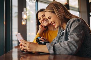 Two women laughing at something on an iPhone while sitting at a café counter