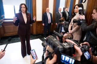 Kamala Harris wearing a maroon suit speaking to a group of press photographers and reporters