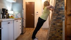 A woman performs wall push-ups in a kitchen. Her hands are on a wall, elbows bent to the side, legs straight and chest close to the wall. 