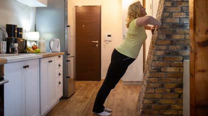 A woman performs wall push-ups in a kitchen. Her hands are on a wall, elbows bent to the side, legs straight and chest close to the wall.