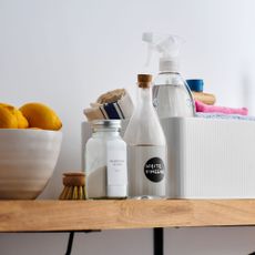 Shelf full of cleaning products, including a bottle of white vinegar, a bowl of lemons, a cleaning caddy, and bicarb