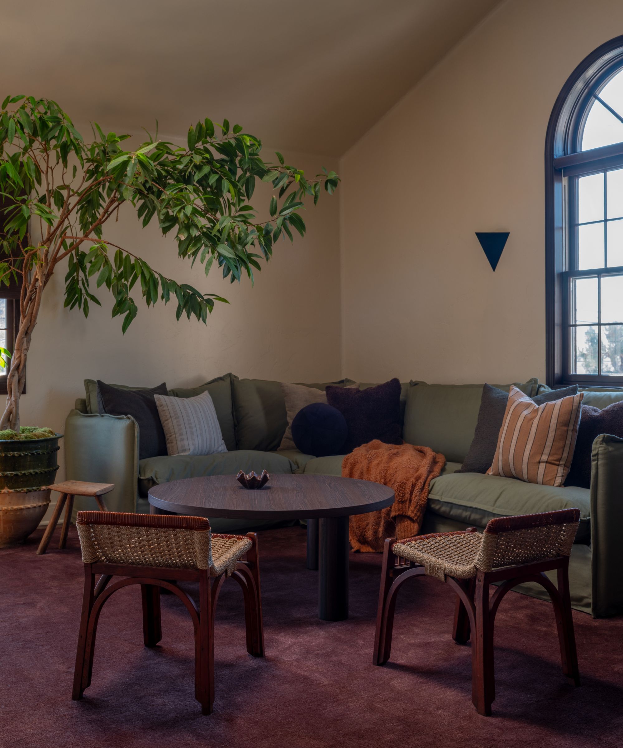 moody living room with a large arched window, large plant, green corner couch, round wooden table and a purple area rug