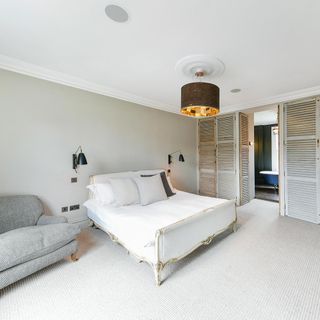 a bedroom with light grey walls and carpet, an elegant yet simple gold and white bed, a grey sofa and wooden shutter doors on the wardrobes and leading to the ensuite bathroom