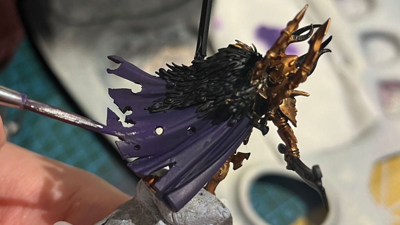 Someone paints a Warhammer model's cloak with a brush loaded with purple paint